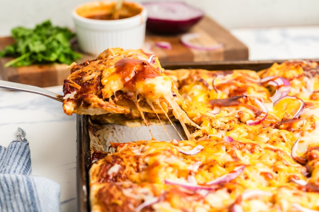 A close-up of a cheesy pizza being lifted from a tray, with melted cheese stretching, red onions on top, and a bowl of sauce, parsley, and a red onion in the background.