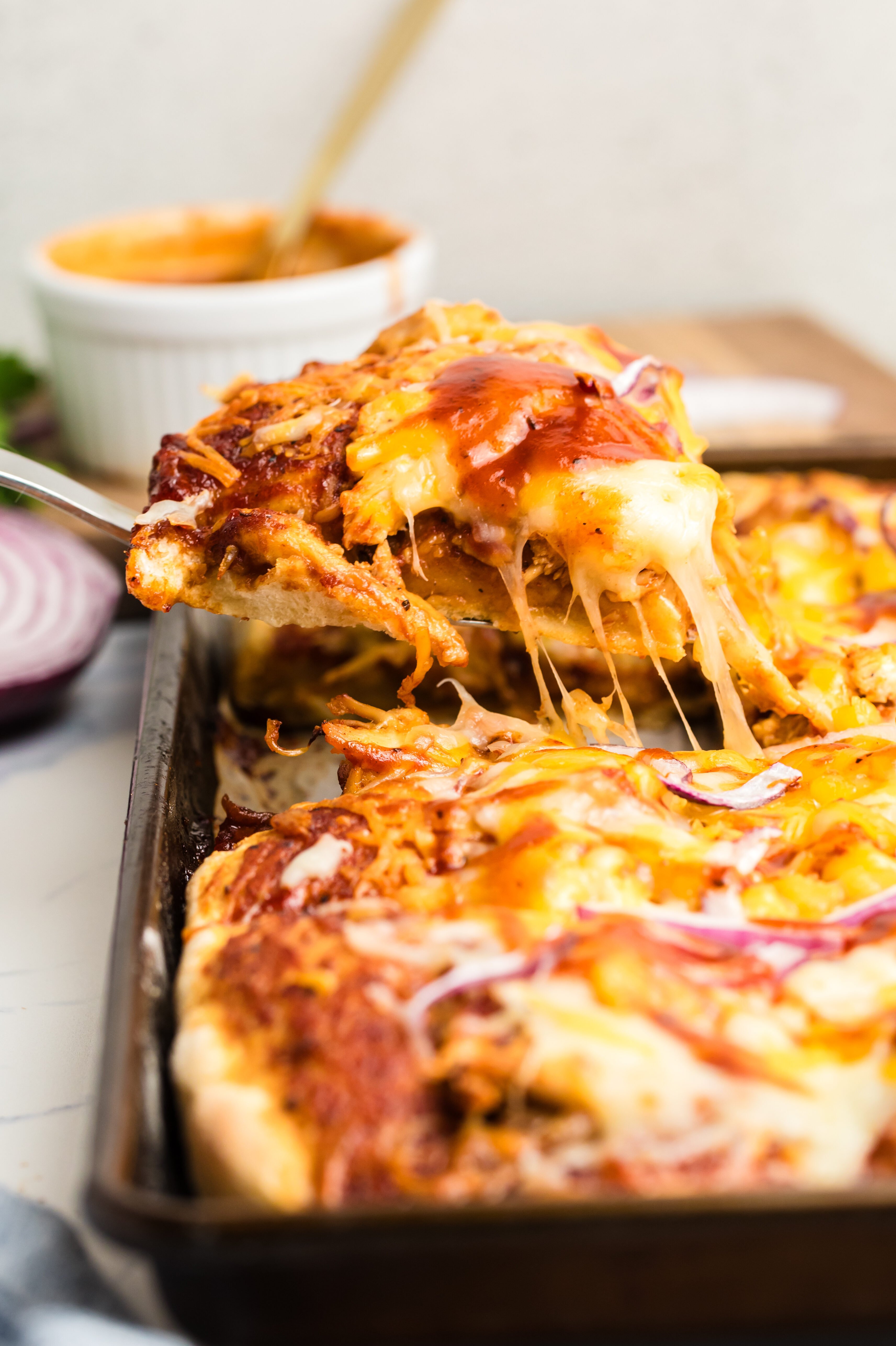 A spatula lifts a cheesy slice of homemade pizza with melted cheese and red onions from a rectangular baking tray. A bowl of sauce and sliced red onion are in the background.