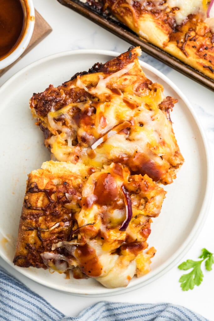 Two slices of cheesy barbecue pizza with red onion on a white plate, with a drizzle of barbecue sauce on top. A baking tray with more pizza and a small bowl of sauce are nearby.