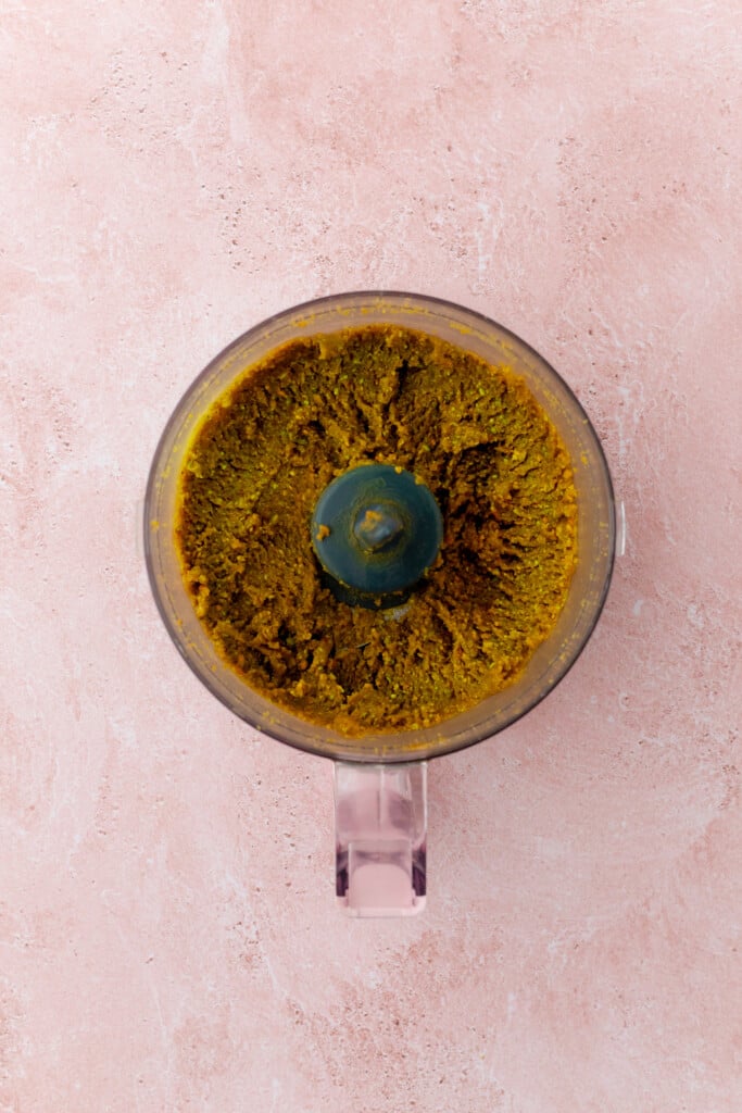 A food processor filled with a coarse, brownish-green paste, viewed from above, sits on a light pink textured surface.