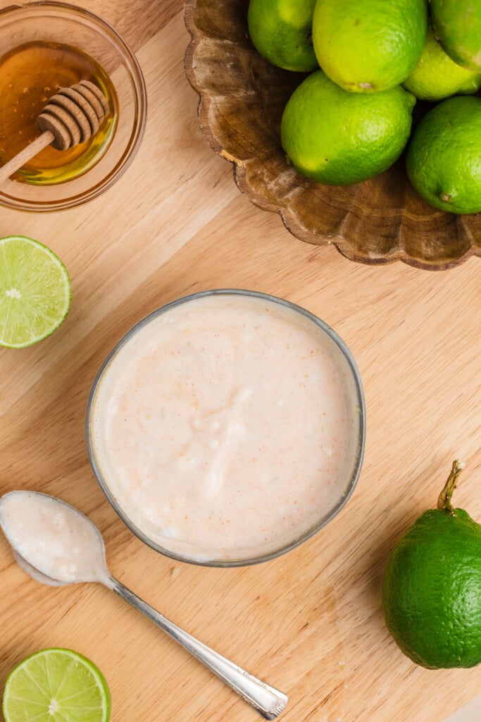 A bowl of creamy sauce on a wooden surface, surrounded by whole and halved limes, a spoon with sauce, and a small dish of honey with a honey dipper.