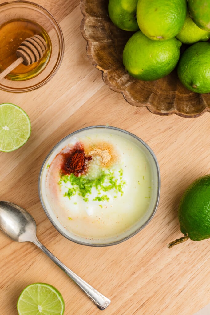 A bowl of yogurt topped with lime zest, ground spices, and chili powder sits on a wooden surface with whole and sliced limes, a spoon, and a small bowl of honey nearby.