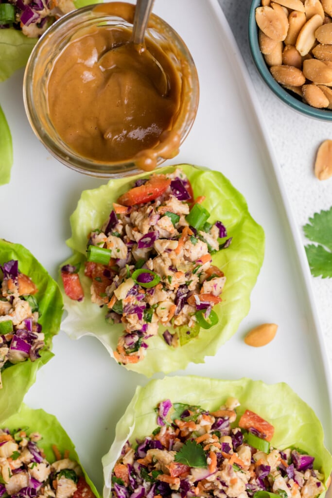 Three lettuce cups filled with a colorful chicken and vegetable mixture are arranged on a white plate. A jar of peanut sauce with a spoon and a bowl of peanuts are nearby. The dish appears fresh and vibrant.