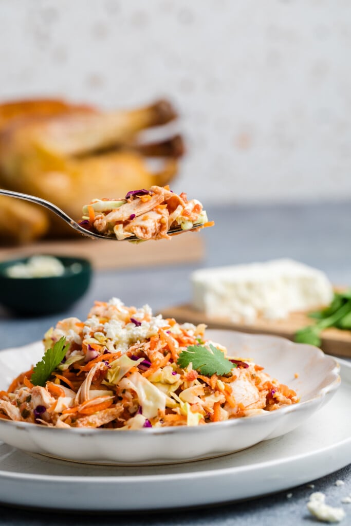 A close-up of a fork holding a bite of Salsa Chicken Coleslaw above a plate. In the background, there is a roasted chicken, a block of cheese, and fresh herbs on a cutting board.