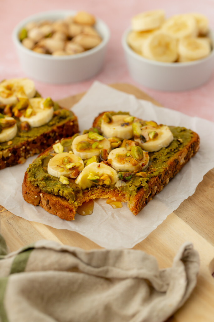A slice of toast topped with green pistachio spread, banana slices, chopped pistachios, and honey sits on parchment paper. In the background are two bowls, one with banana slices and the other with pistachios.