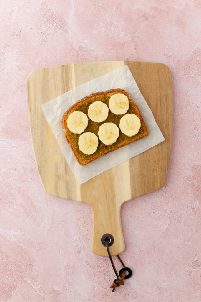 A slice of toast topped with six banana slices sits on parchment paper on a wooden cutting board, placed on a pink textured surface.