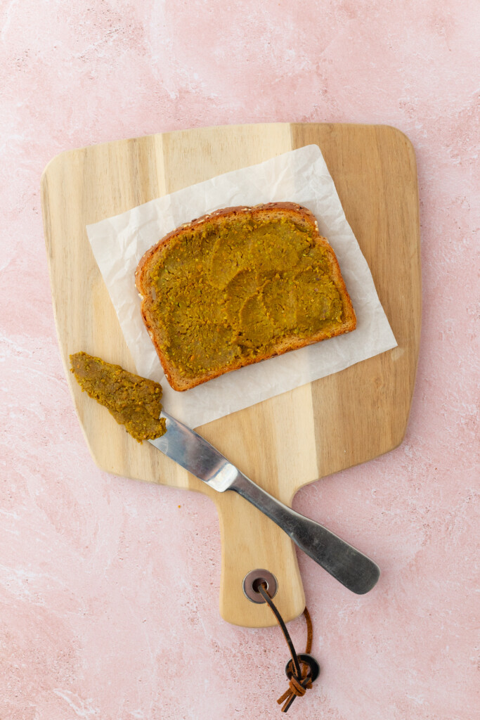 A slice of bread with greenish-brown spread on parchment paper atop a wooden cutting board, next to a knife with more spread, on a pink textured surface.
