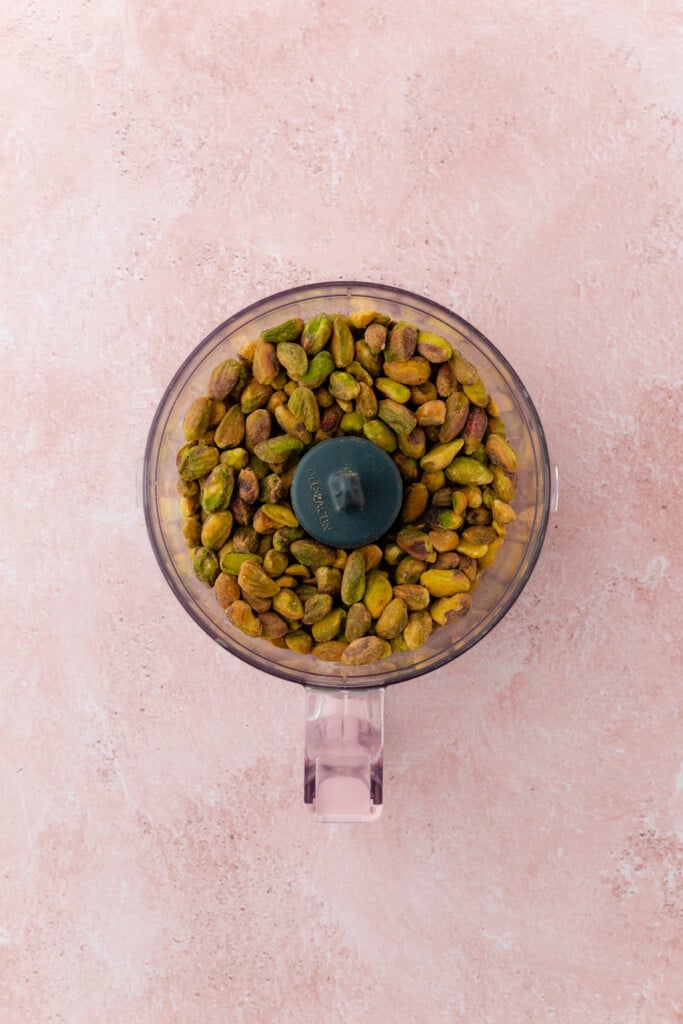 A food processor bowl filled with whole shelled pistachios sits on a pink textured surface, viewed from above.