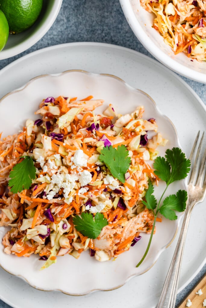 A plate of colorful Salsa Chicken Coleslaw with carrots, red cabbage, cheese crumbles, and fresh cilantro, served on a white dish with a fork and knife beside it. A bowl of limes and extra slaw are nearby.