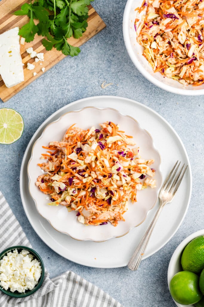 A plate of vibrant Salsa Chicken Coleslaw made with shredded cabbage and carrots sits on a white plate with a fork. Surrounding items include fresh cilantro, feta cheese, lime halves, and a bowl of extra coleslaw.