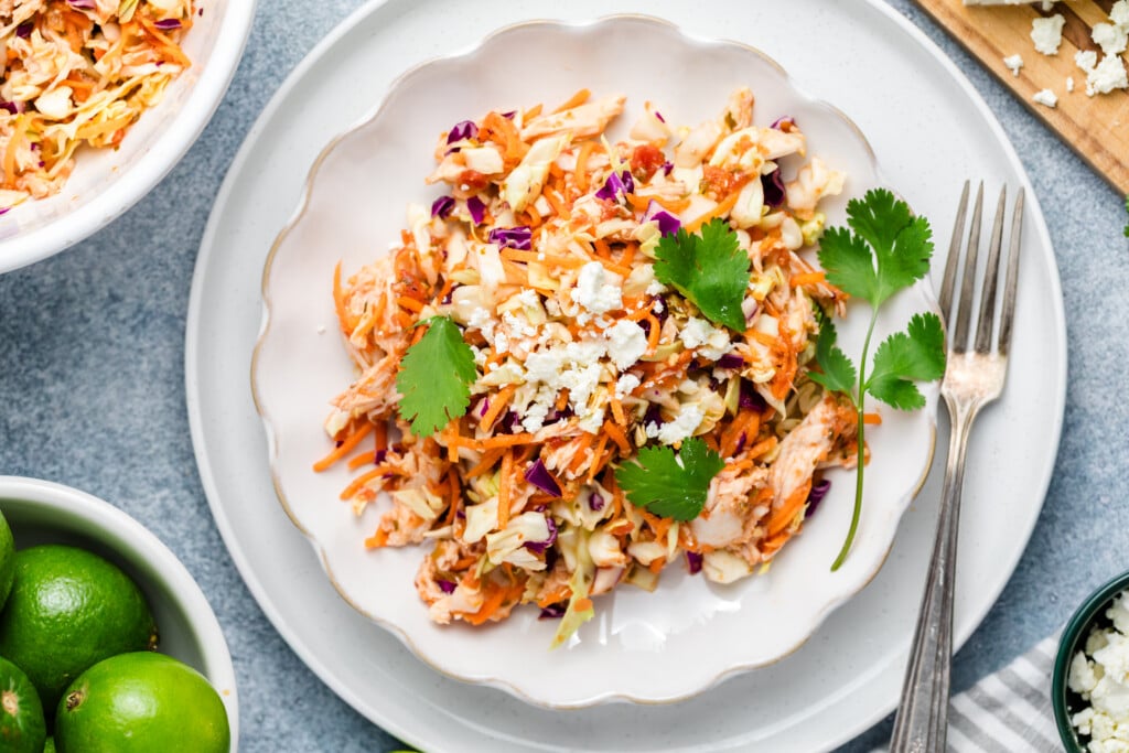 A plate of salsa chicken coleslaw with shredded cabbage, carrots and chicken, garnished with cilantro and crumbled cheese, placed on a white plate with a fork. A bowl of lemon and extra lettuce is nearby.