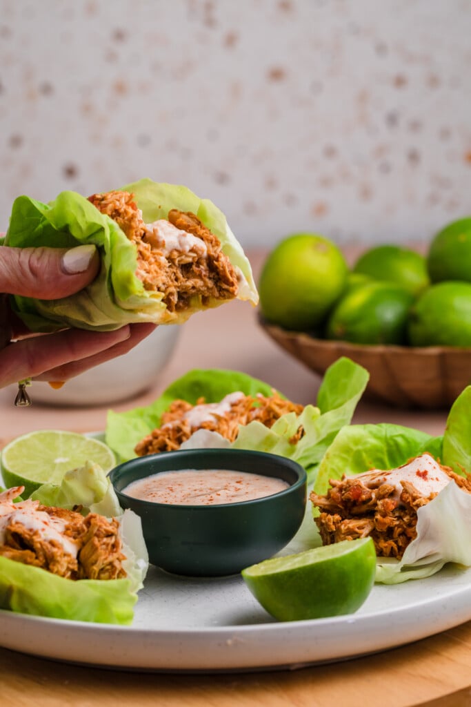 A hand holds a lettuce wrap filled with shredded spiced chicken and sauce. Other lettuce wraps, lime wedges, and a bowl of dipping sauce are on the table, with a bowl of whole limes in the background.