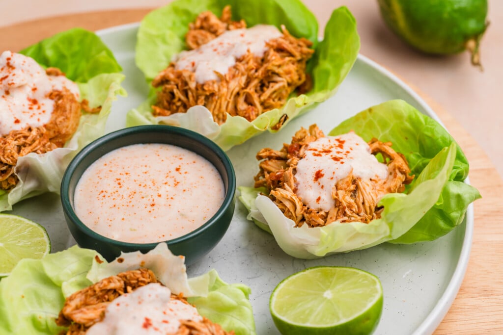 A plate of lettuce wraps is filled with shredded chicken and topped with a creamy sauce, with a bowl of dipping sauce and lemon halves on the side.