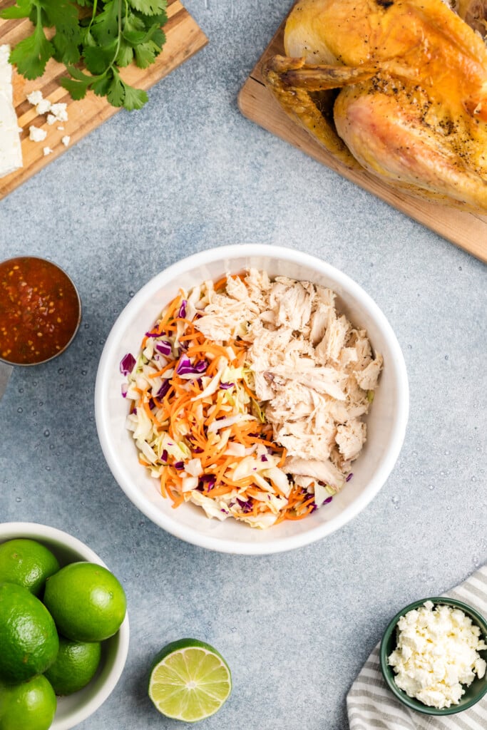 A white bowl with shredded Salsa Chicken and mixed coleslaw sits on a gray surface, surrounded by a roast chicken, fresh limes, zesty salsa, cilantro, and cheese.