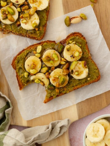 Two slices of toast topped with green pistachio spread, banana slices, chopped pistachios, and a drizzle of honey, arranged on parchment paper. One toast has a bite taken out. A bowl of banana slices is nearby.