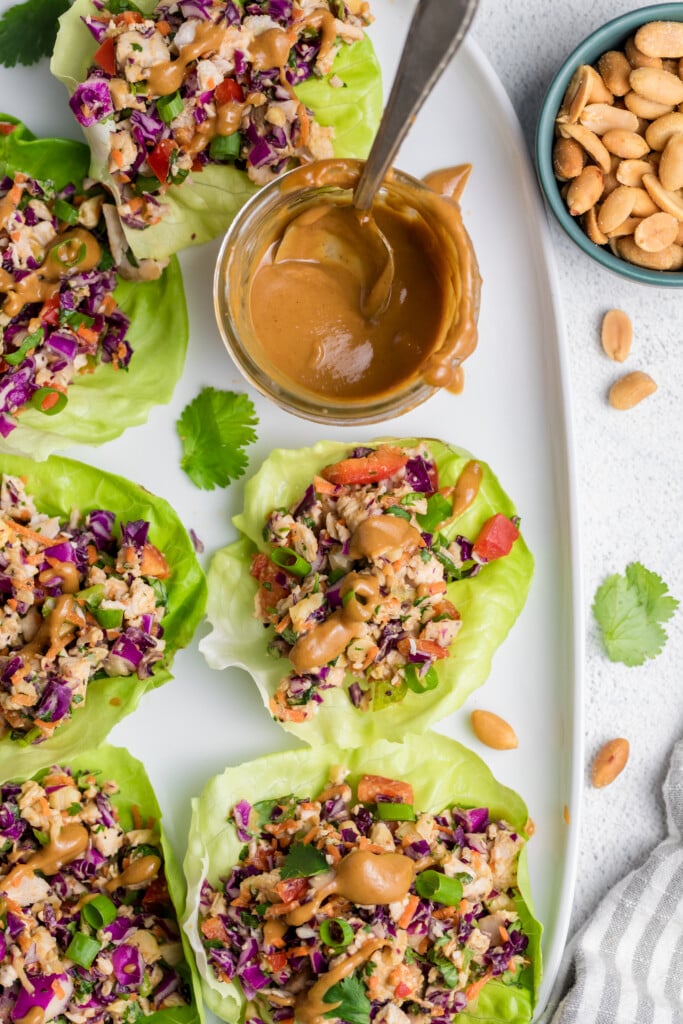 A platter of lettuce wraps filled with colorful chopped vegetables and peanuts, drizzled with a creamy peanut sauce. A jar of sauce with a spoon and a bowl of peanuts are placed nearby for serving.