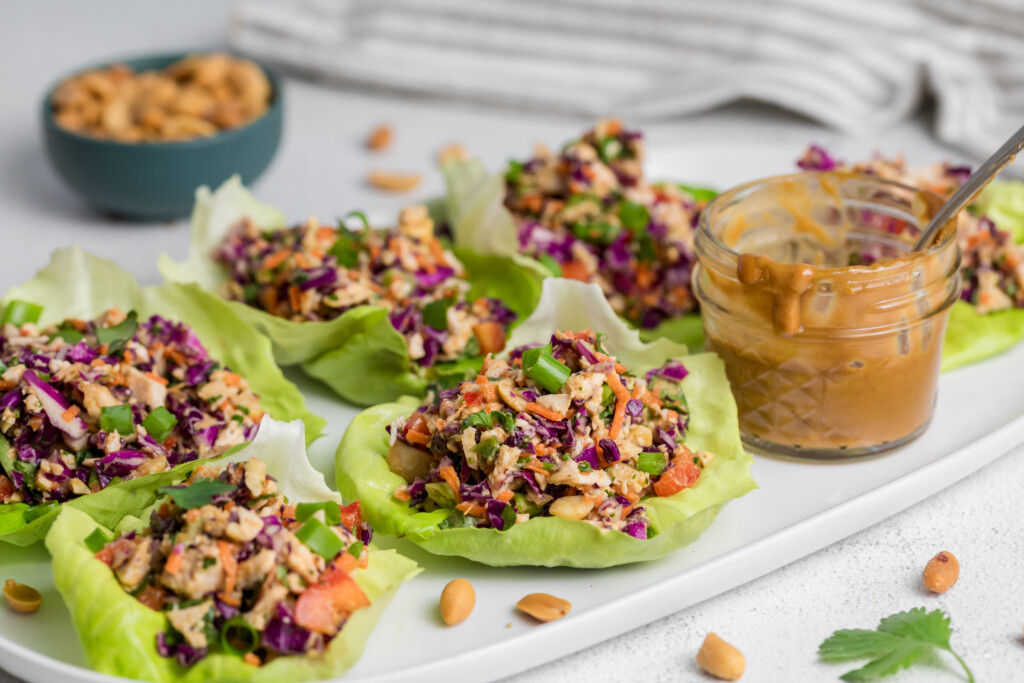 Six lettuce cups filled with a colorful mix of chopped vegetables, herbs, and peanuts are arranged on a white tray. A small glass jar of creamy peanut sauce sits beside them, with a few peanuts scattered nearby.