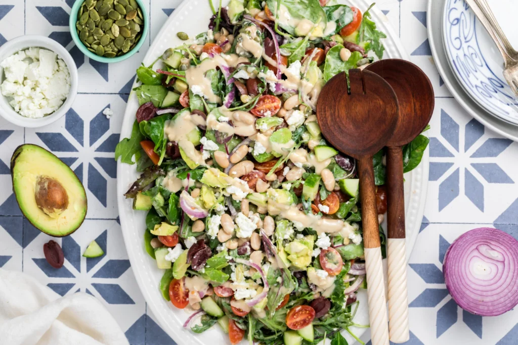 A large oval platter of mixed green salad with cherry tomatoes, cucumber, beans, red onion, feta cheese, olives, and avocado, topped with creamy dressing. Wooden salad servers and small bowls of toppings are nearby.