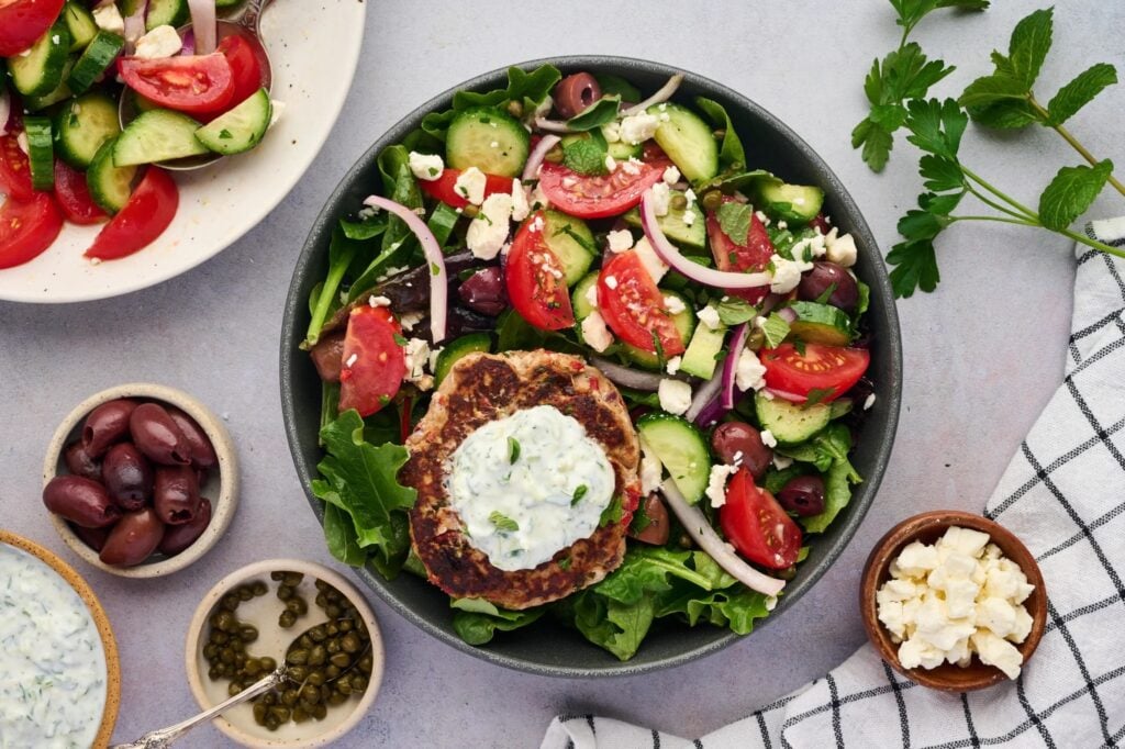 A bowl of Greek salad with cucumbers, tomatoes, olives, red onions, feta and greens topped with a chicken patty and tzatziki sauce – perfect for a high-protein lunch. Bowls of olives, capers, feta, herbs and a cloth napkin are nearby.