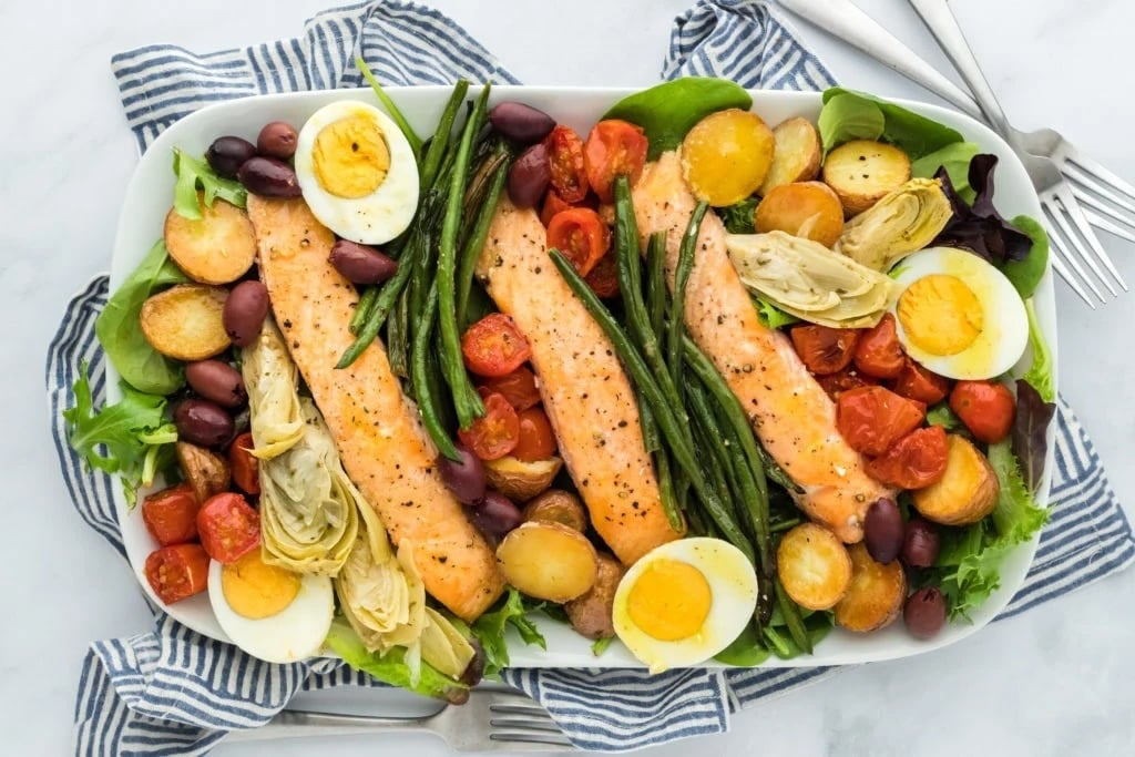 A platter of baked salmon fillets, green beans, roasted potatoes, cherry tomatoes, olives, artichokes, hard-boiled egg halves and mixed green vegetables arranged neatly on a striped towel – perfect for a high-protein lunch.