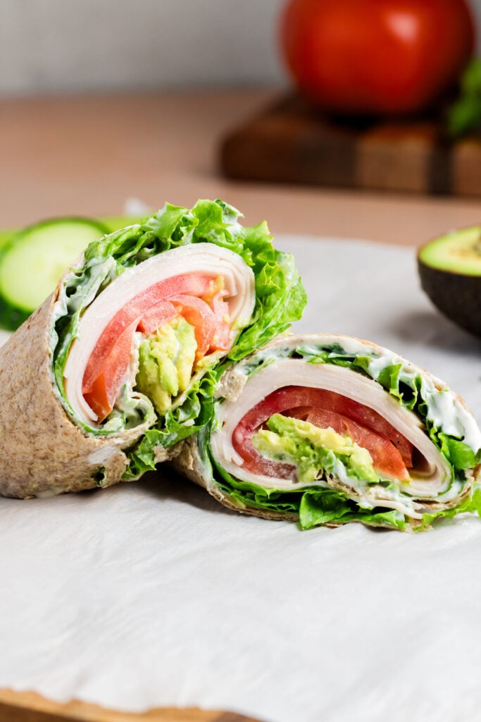 A California Turkey Wrap, sliced in half and filled with lettuce, tomato, avocado, and a creamy spread, sits on parchment paper. Sliced cucumbers, half an avocado, and a tomato on a cutting board are in the background.