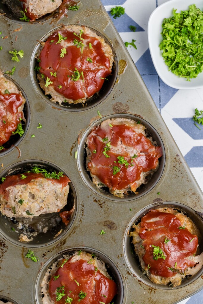 A muffin tin filled with Mini Meatloaf Muffins, each topped with ketchup and garnished with chopped parsley, sits alongside a small bowl of fresh parsley on a patterned surface.