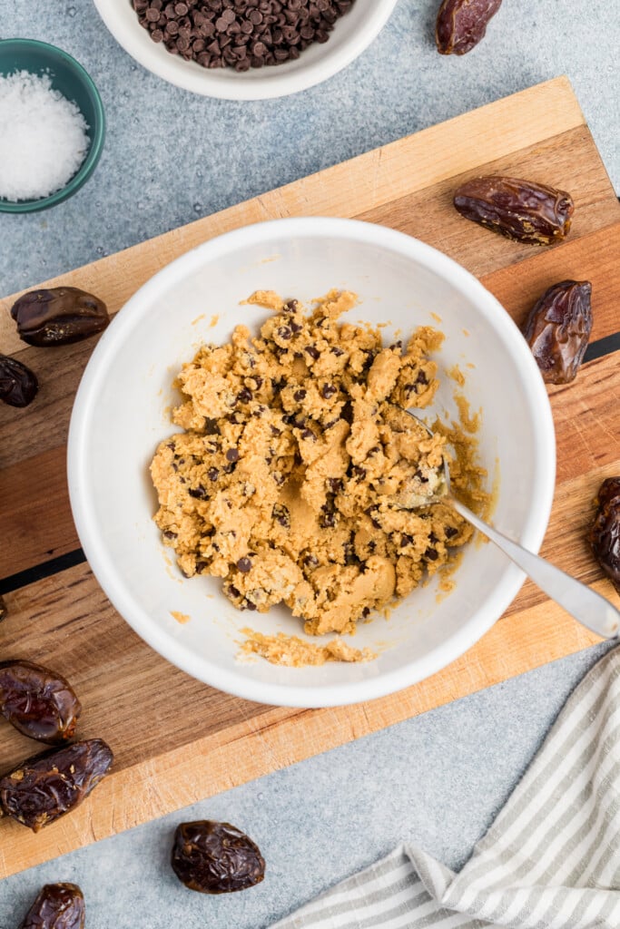 A white bowl of chocolate chip cookie dough with a spoon on a wooden cutting board, surrounded by Medjool dates ready for Chocolate Chip Cookie Dough Stuffed Dates, plus a bowl of chocolate chips and a small dish of flaky sea salt.