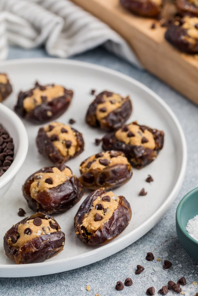 A plate of Chocolate Chip Cookie Dough Stuffed Dates, filled with peanut butter and mini chocolate chips, sits beside a bowl of chocolate chips and a pinch bowl of salt. More stuffed dates rest in the background on a wooden tray.
