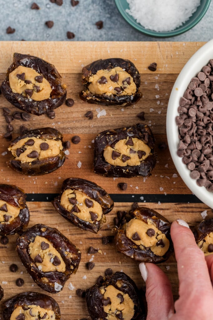 Chocolate Chip Cookie Dough Stuffed Dates filled with peanut butter and topped with mini chocolate chips are arranged on a wooden board. A hand reaches for one, while bowls of mini chocolate chips and flaky sea salt sit nearby.