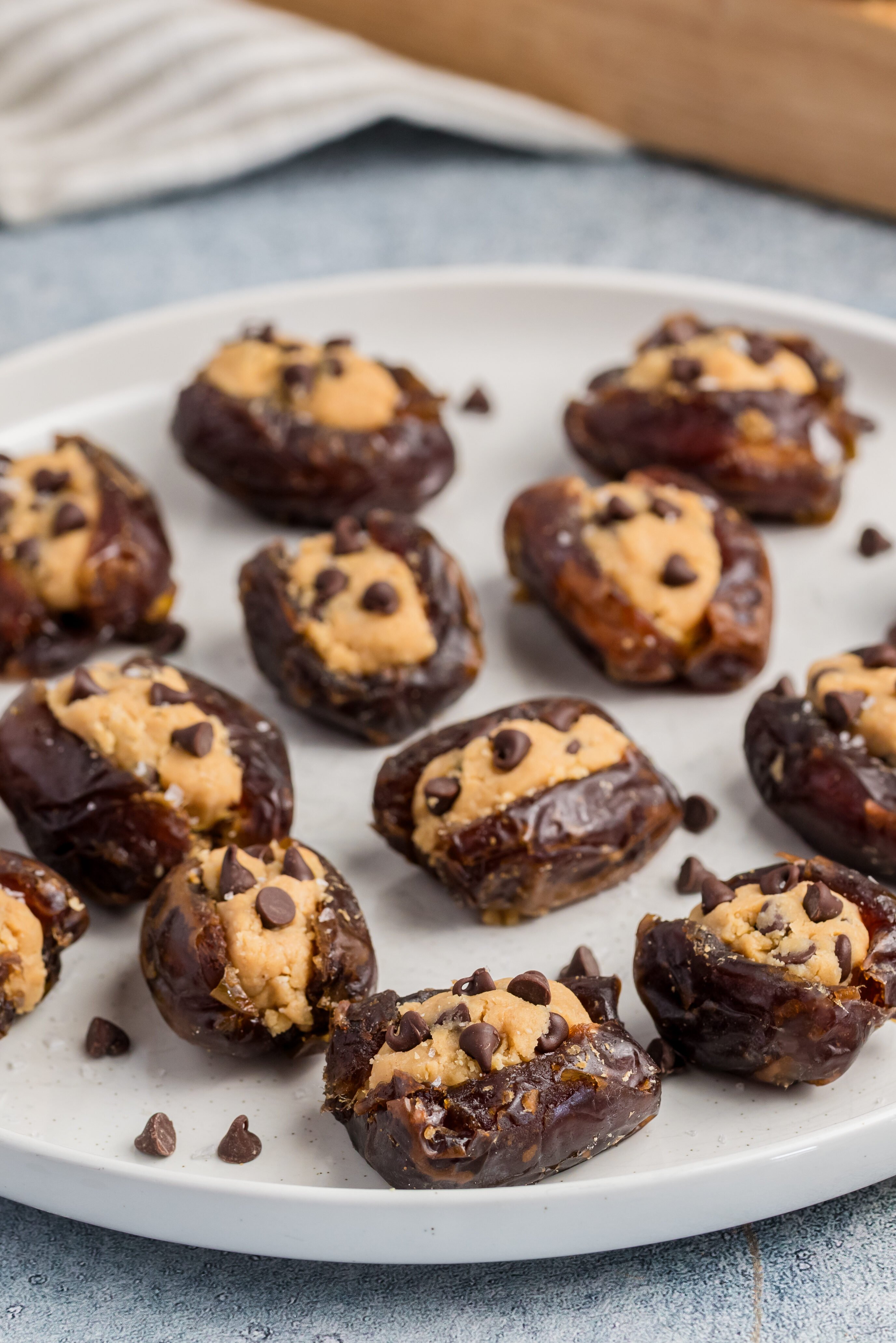 A white plate filled with Chocolate Chip Cookie Dough Stuffed Dates, brimming with creamy filling and topped with mini chocolate chips. More dates, chocolate chips, and a bowl of salt surround the plate on a light surface.