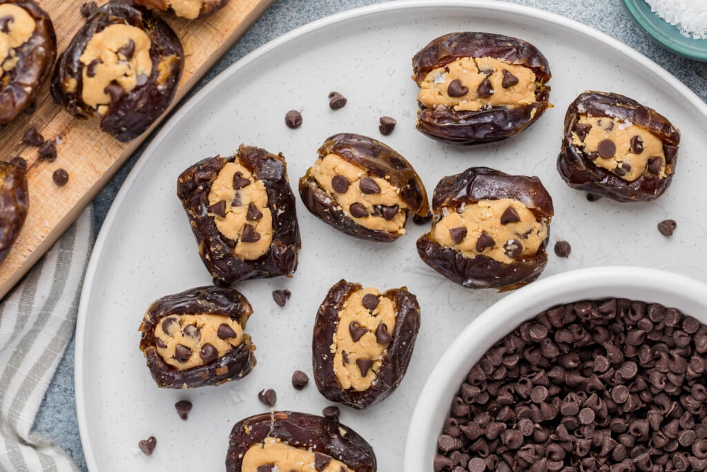 A plate of Chocolate Chip Cookie Dough Stuffed Dates, filled with peanut butter and topped with mini chocolate chips, sits alongside a bowl of chocolate chips and more stuffed dates on a wooden board.