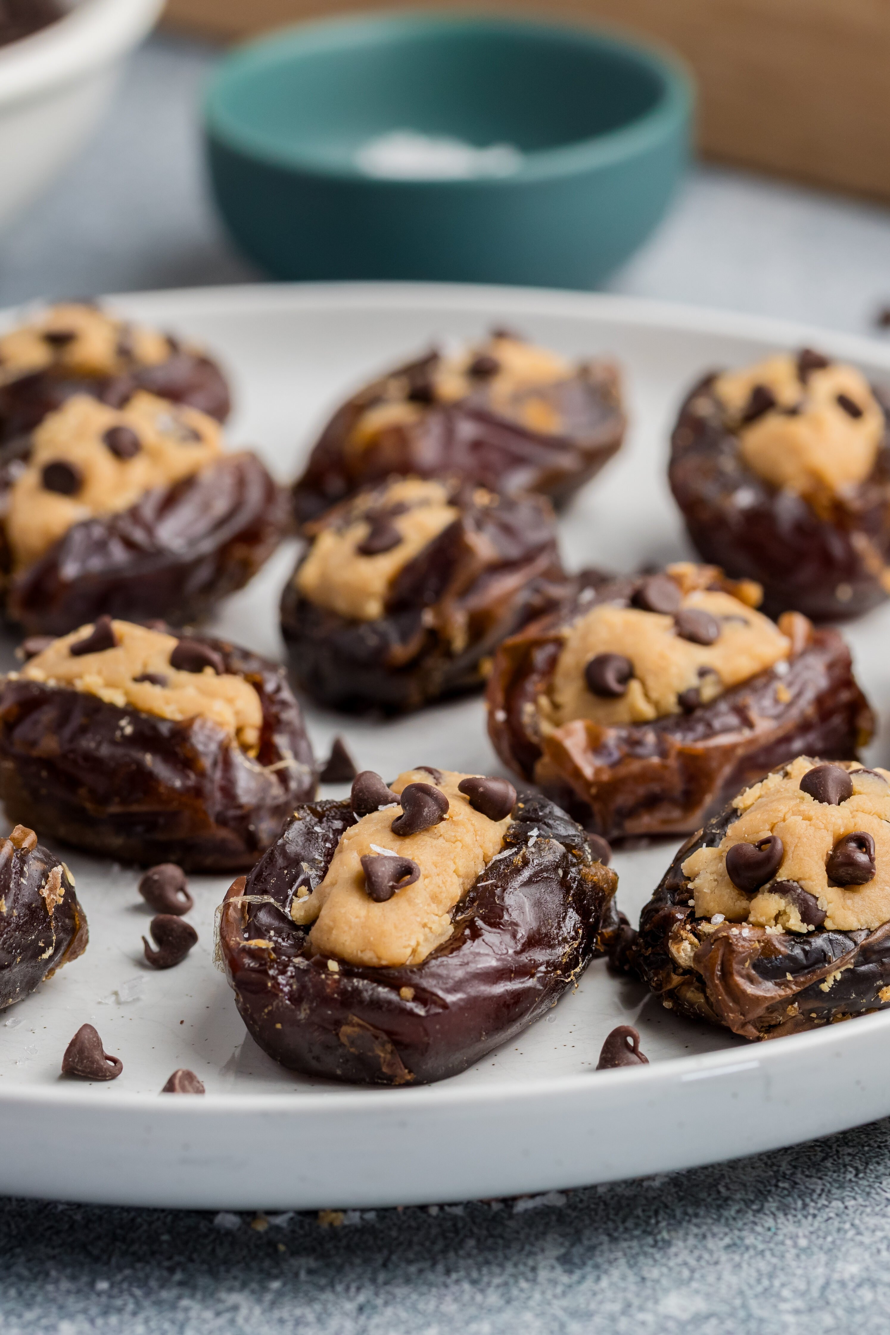 A plate of Chocolate Chip Cookie Dough Stuffed Dates, filled with nut butter and topped with mini chocolate chips, arranged on a round white plate with extra chocolate chips scattered around.