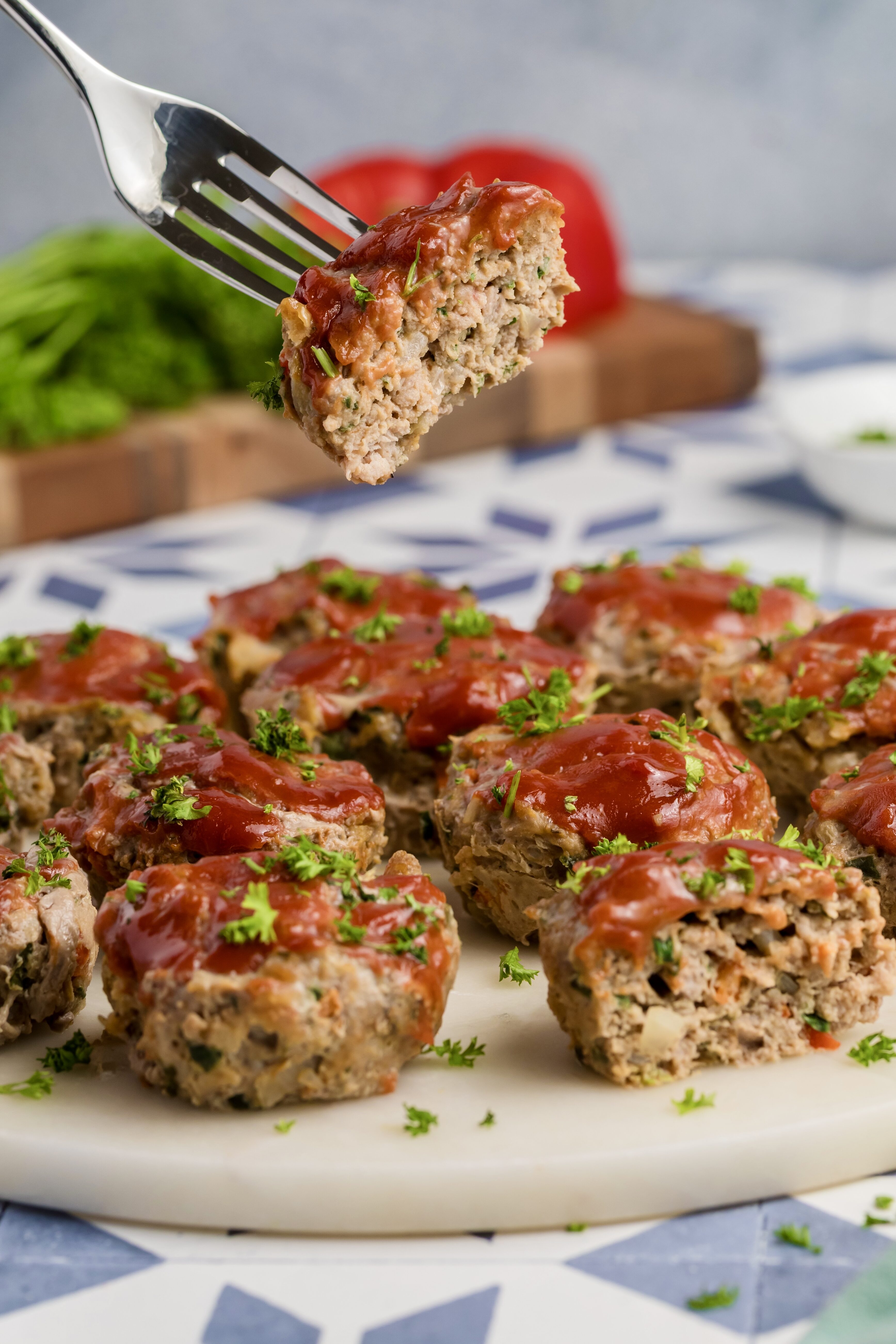 A fork holds up a Mini Meatloaf Muffin topped with ketchup glaze, with several more mini meatloaf muffins arranged on a white plate, garnished with chopped parsley. A blurred background shows herbs and a red bell pepper.