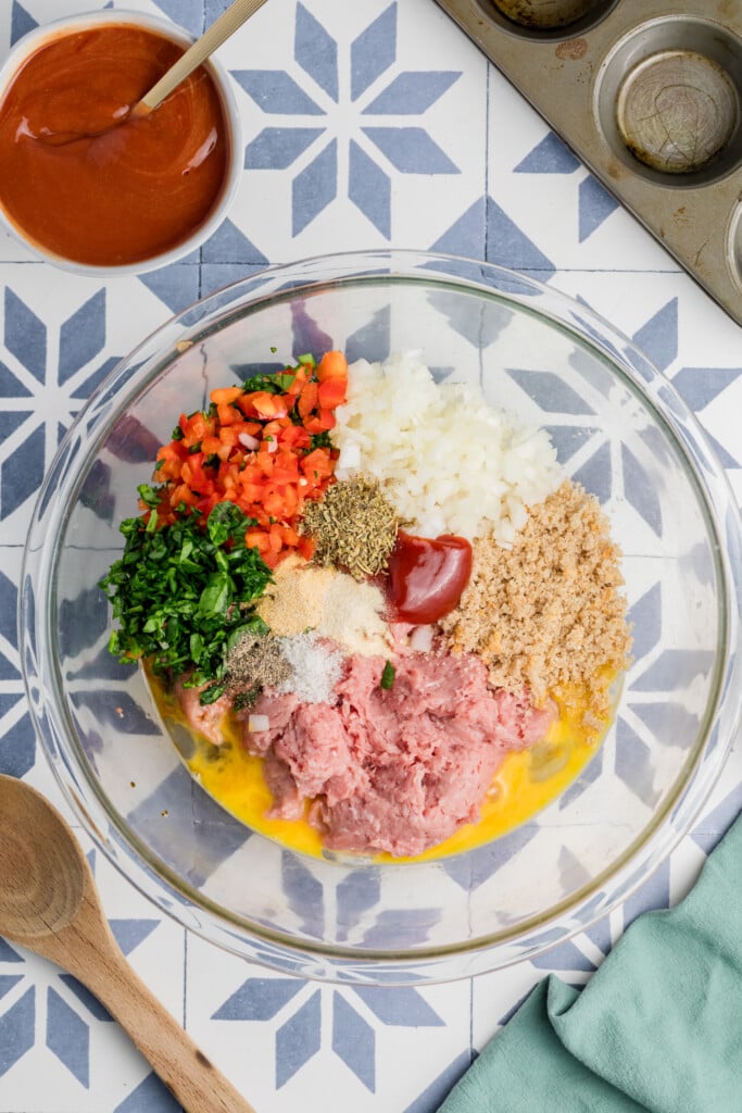 A glass bowl holds the ingredients for Mini Meatloaf Muffins—ground meat, chopped onions, red bell pepper, parsley, breadcrumbs, ketchup, seasonings, and eggs. Nearby are a bowl of sauce, a muffin tin, and a wooden spoon on a patterned surface.
