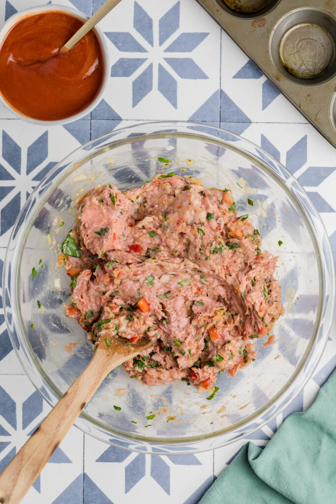 A glass bowl with raw ground meat mixed with herbs, red pepper, and onion is stirred for Mini Meatloaf Muffins. A bowl of tomato sauce and a muffin tin sit nearby on a blue patterned surface.