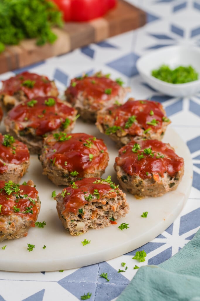 Mini Meatloaf Muffins topped with ketchup glaze and garnished with chopped parsley are arranged on a round marble platter, with extra parsley in a small bowl nearby. The background features a blue and white geometric pattern.