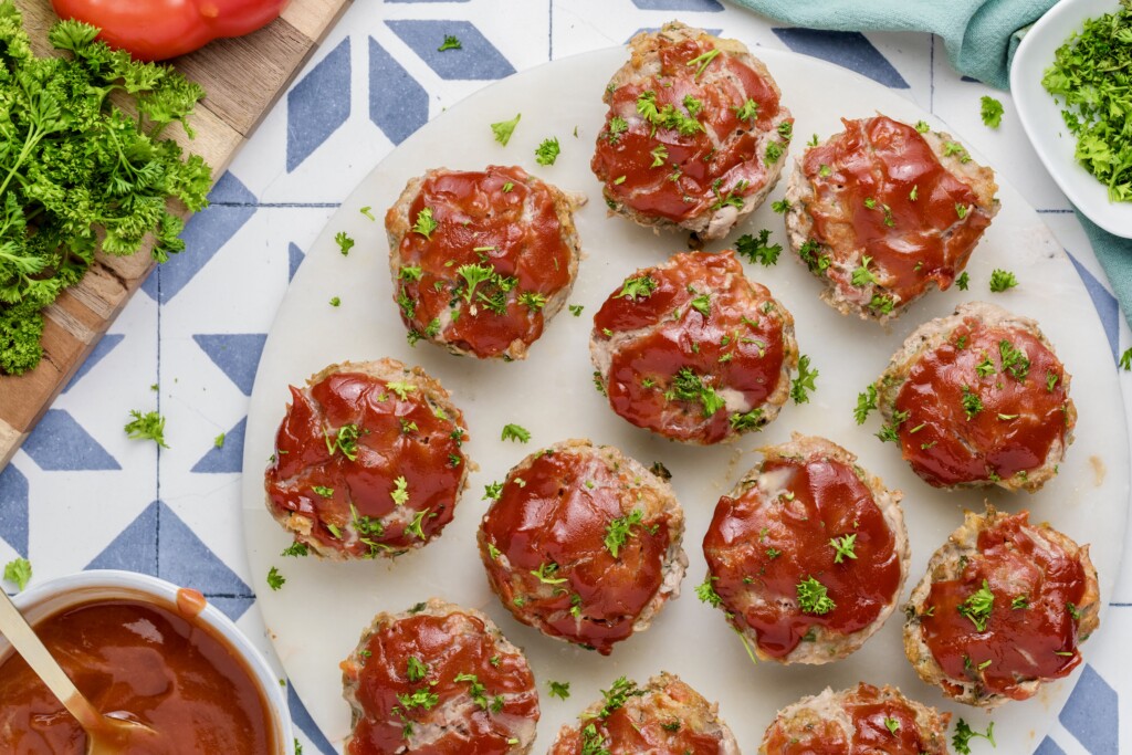 A round white plate with Mini Meatloaf Muffins topped with red glaze and sprinkled with chopped parsley, surrounded by a bowl of sauce, fresh parsley, and a blue patterned surface.