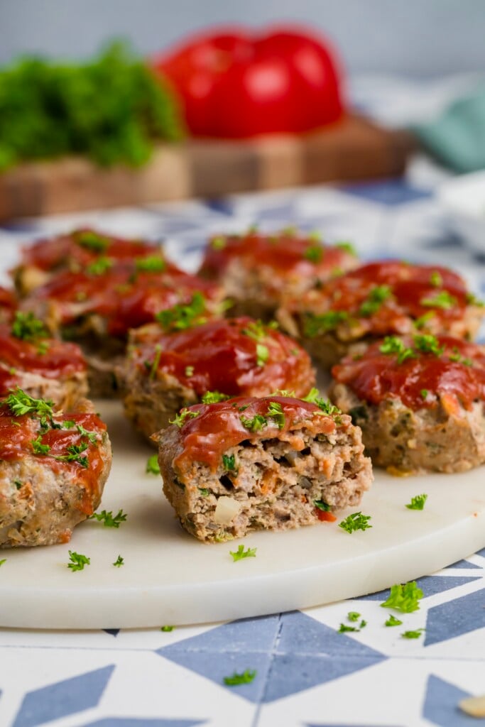 Mini Meatloaf Muffins topped with tomato sauce and chopped parsley sit on a round white platter, with one showing a bite revealing the inside. Fresh vegetables are blurred in the background.