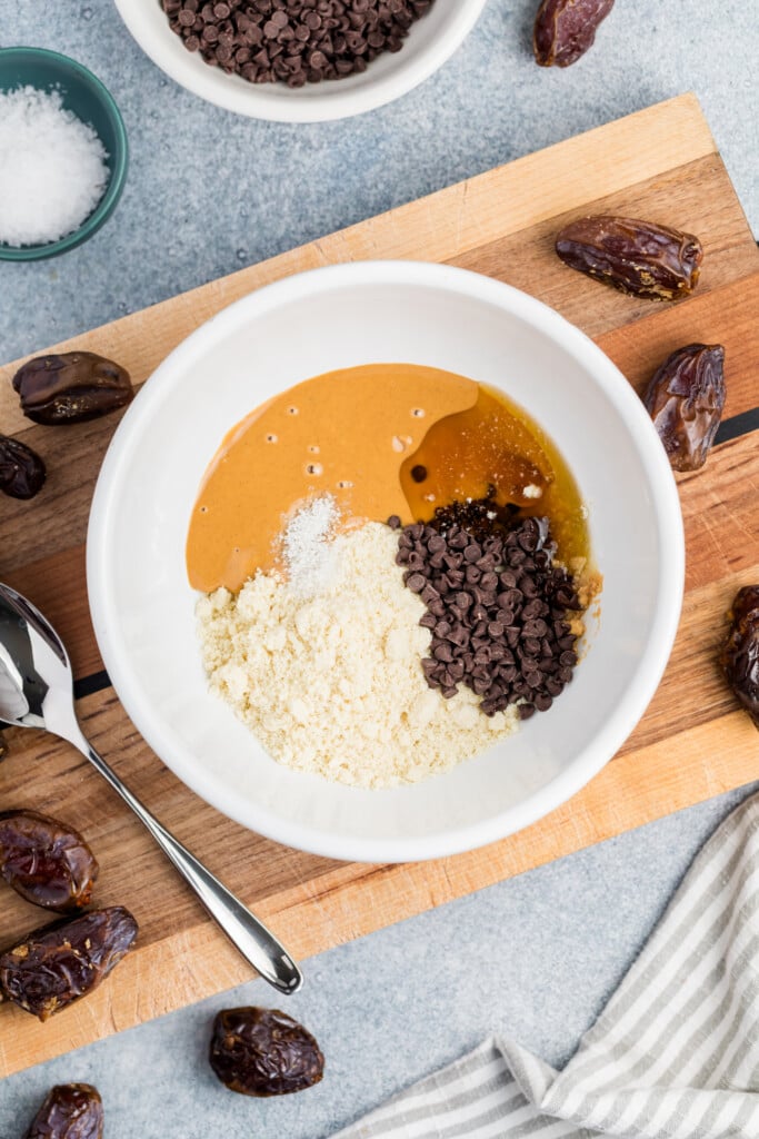 A white bowl on a wooden board holds ingredients for Chocolate Chip Cookie Dough Stuffed Dates: peanut butter, almond flour, chocolate chips, honey, and salt. Nearby are dates and a spoon, with a striped cloth and small bowl of salt in the background.