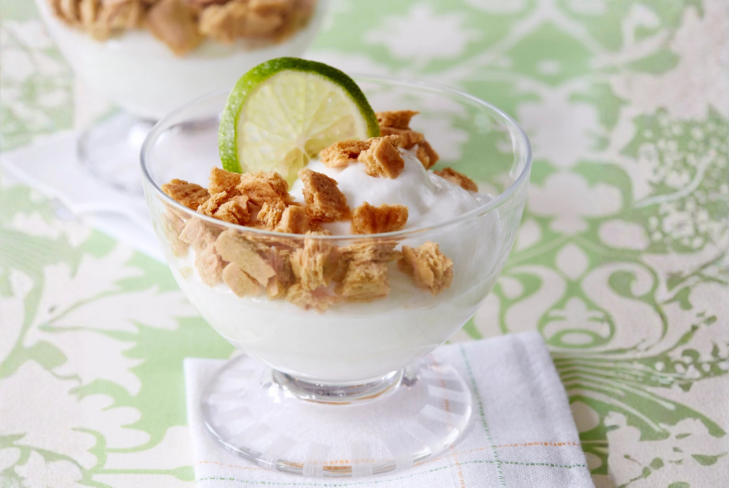 A glass bowl filled with yogurt, one of the best high protein snacks, topped with crumbled cereal and garnished with a slice of lime, sits on a white napkin over a green and white patterned surface.