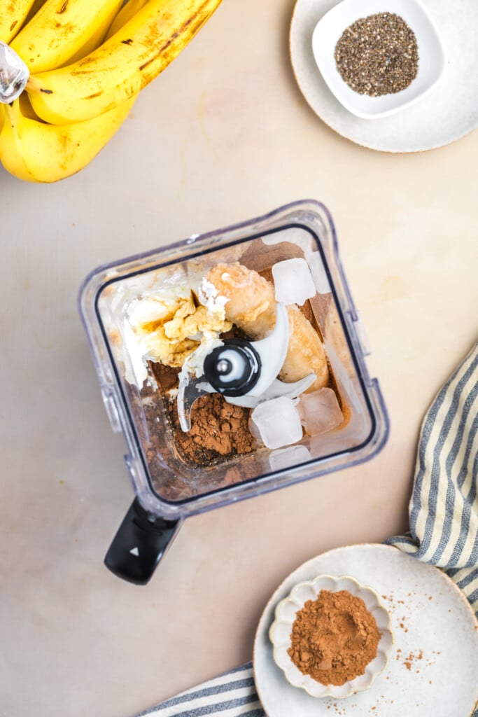 Top-down view of a blender containing ice cubes, banana, yogurt, and cocoa powder—perfect for a Healthy High-Protein Chocolate Smoothie—surrounded by bananas, chia seeds, and cocoa powder on a light surface. No added sugar required!.