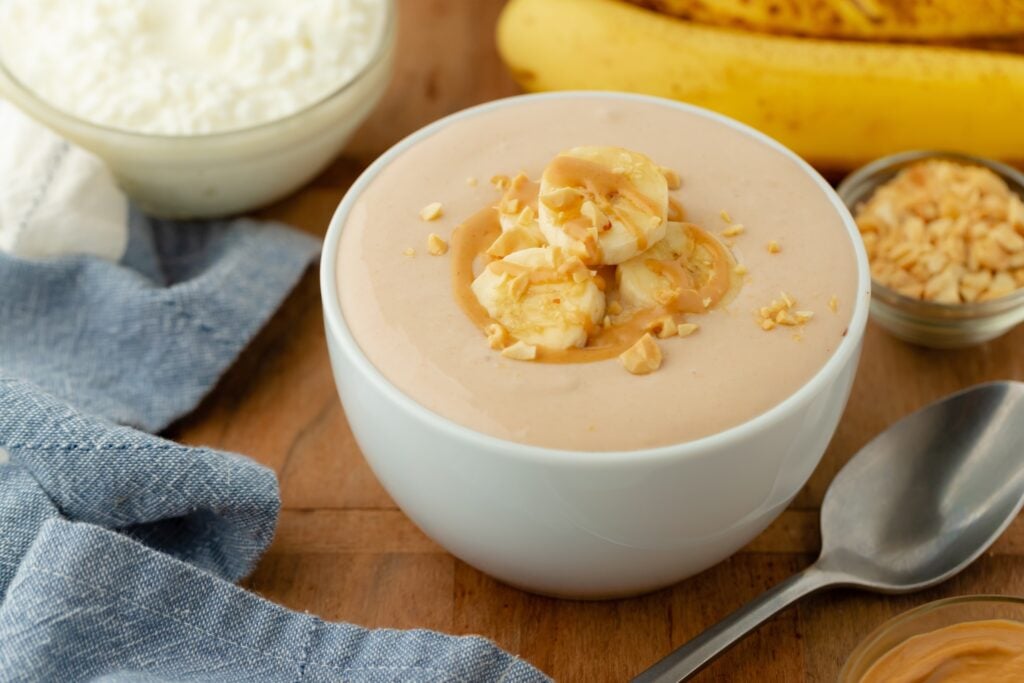 A bowl of creamy peanut butter banana yogurt, a delicious high protein snack, topped with banana slices, chopped peanuts, and a peanut butter drizzle. Nearby are a spoon, blue cloth, cottage cheese, bananas, and peanuts on a wooden table.