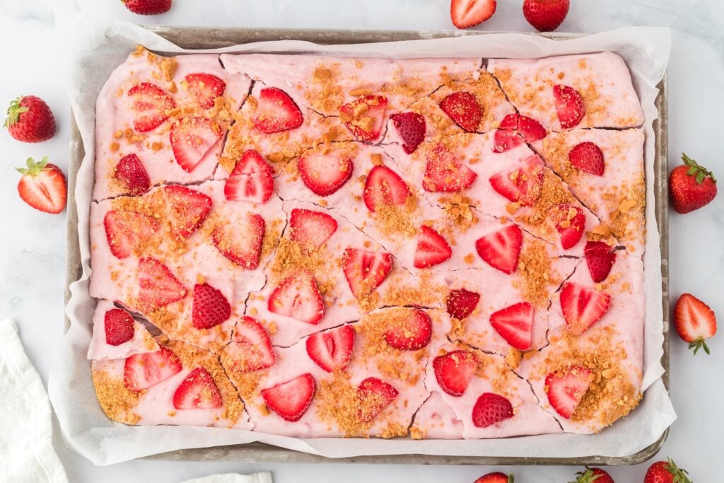 A sheet pan of high protein strawberry dessert bars with a pink creamy topping, fresh strawberry slices, and a crumbly golden topping, set on parchment paper with whole strawberries scattered around. Perfect for high protein snacks.