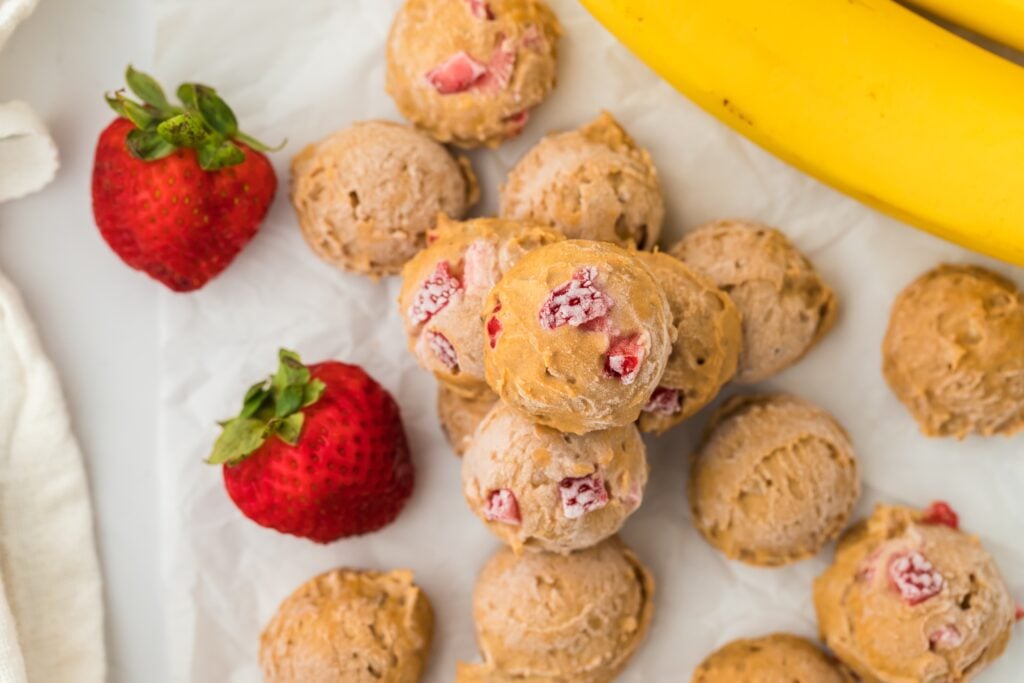 Scoops of strawberry banana ice cream—one of our favorite high protein snacks—are arranged on parchment paper, surrounded by fresh strawberries and whole bananas. The creamy texture features visible pieces of strawberry.