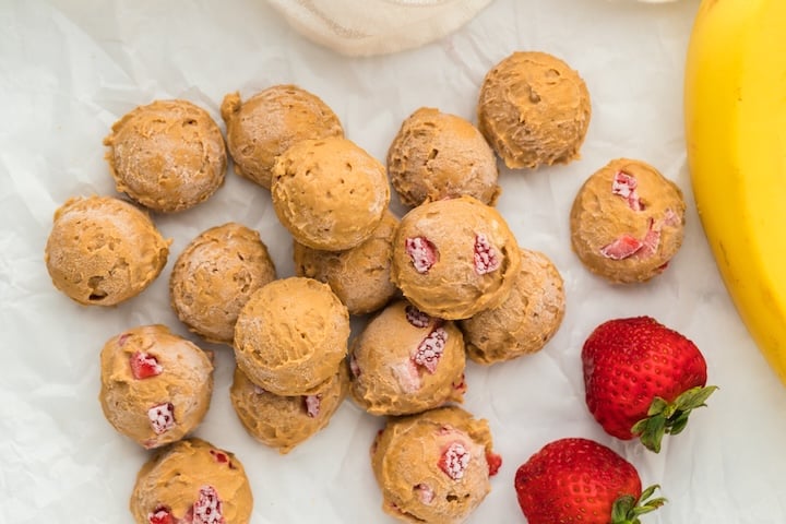 Scoops of light brown, high protein dough with pieces of red strawberries are arranged on white parchment paper, surrounded by fresh strawberries and part of a banana.