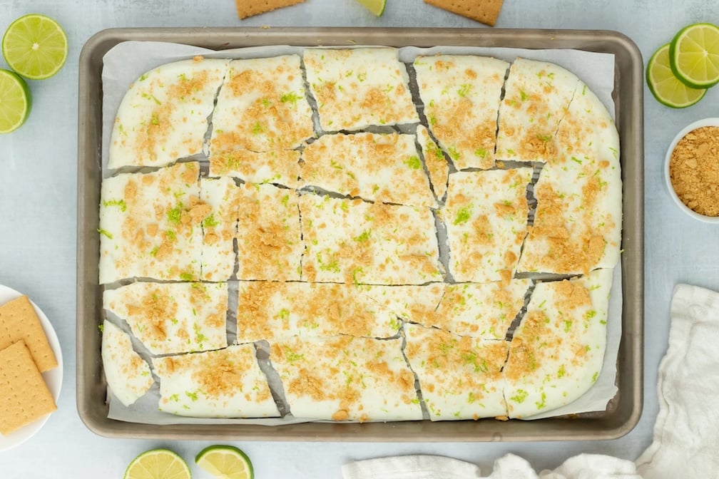 A baking tray with creamy, high protein snack bars topped with lime zest and crushed graham crackers, cut into uneven pieces. Lime slices, graham crackers, and a bowl of crumbs are arranged around the tray.