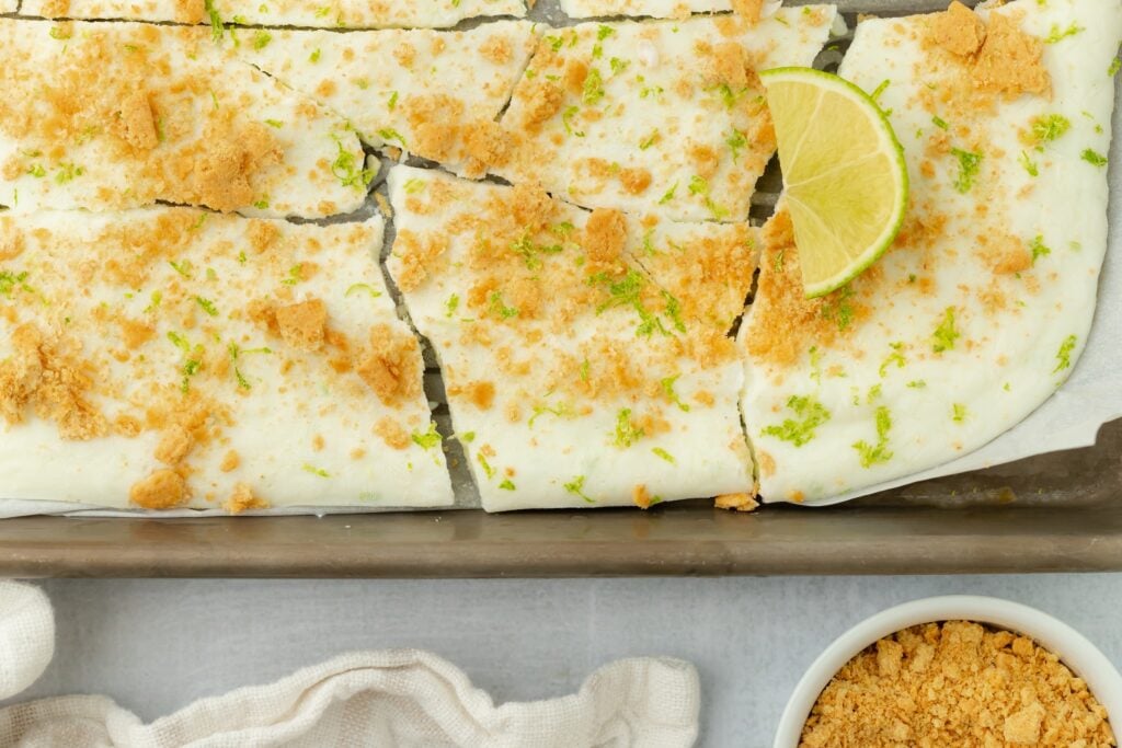 Rectangular dessert with a creamy white topping, sprinkled with graham cracker crumbs and lime zest, cut into squares and garnished with a lime wedge—perfect for those seeking high protein snacks. A bowl of crumbs sits nearby on a light surface.