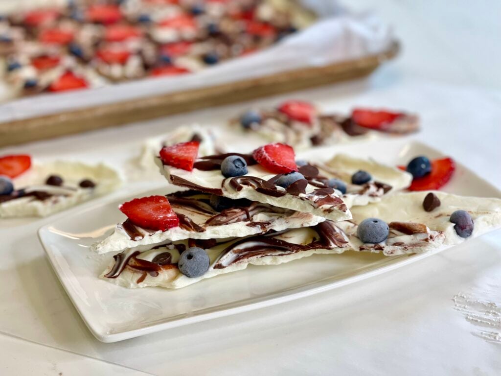 A plate of white yogurt bark topped with strawberries, blueberries, chocolate drizzle, and chocolate chips—one of the best high protein snacks. More pieces are visible on a tray in the background, all served on a white rectangular plate.