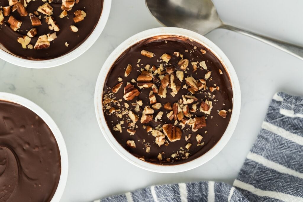 A small bowl of chocolate pudding, perfect for high protein snacks, is topped with chopped pecans and sits on a white surface next to a spoon, a blue and white striped cloth, and two other bowls, one partially visible.