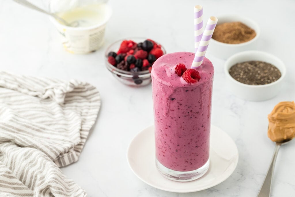 A glass of berry smoothie with two striped straws is topped with raspberries. Surrounding the glass are bowls of mixed berries, chia seeds, flaxseed, yogurt, and a spoonful of peanut butter—perfect high protein snacks on a white surface.